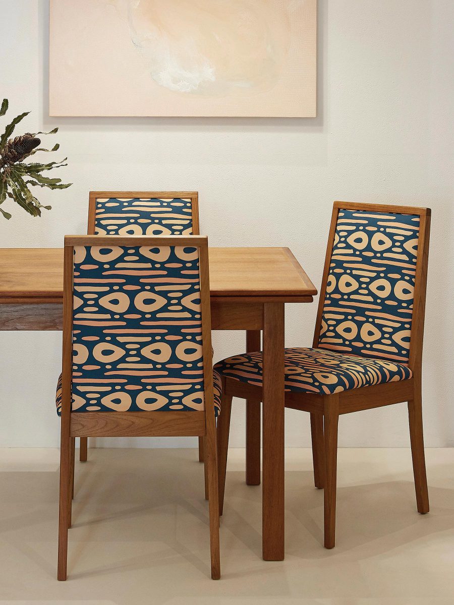 Interior design photo showing part of a wooden dining table and chairs featuring SURFACE 1°22 dot dot dash geometric pattern in teal, clay and lemon colours with abstract painting on the white wall
