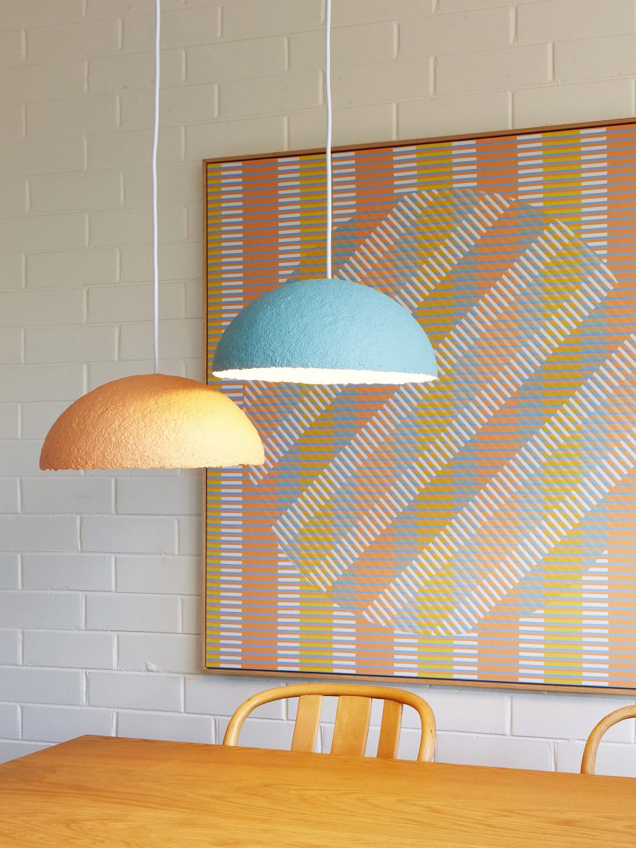 Interior styling against a white brick wall featuring an orange striped pattern wall hanging, two ceiling lamps in orange and blue, the corner of a wooden table and chairs.